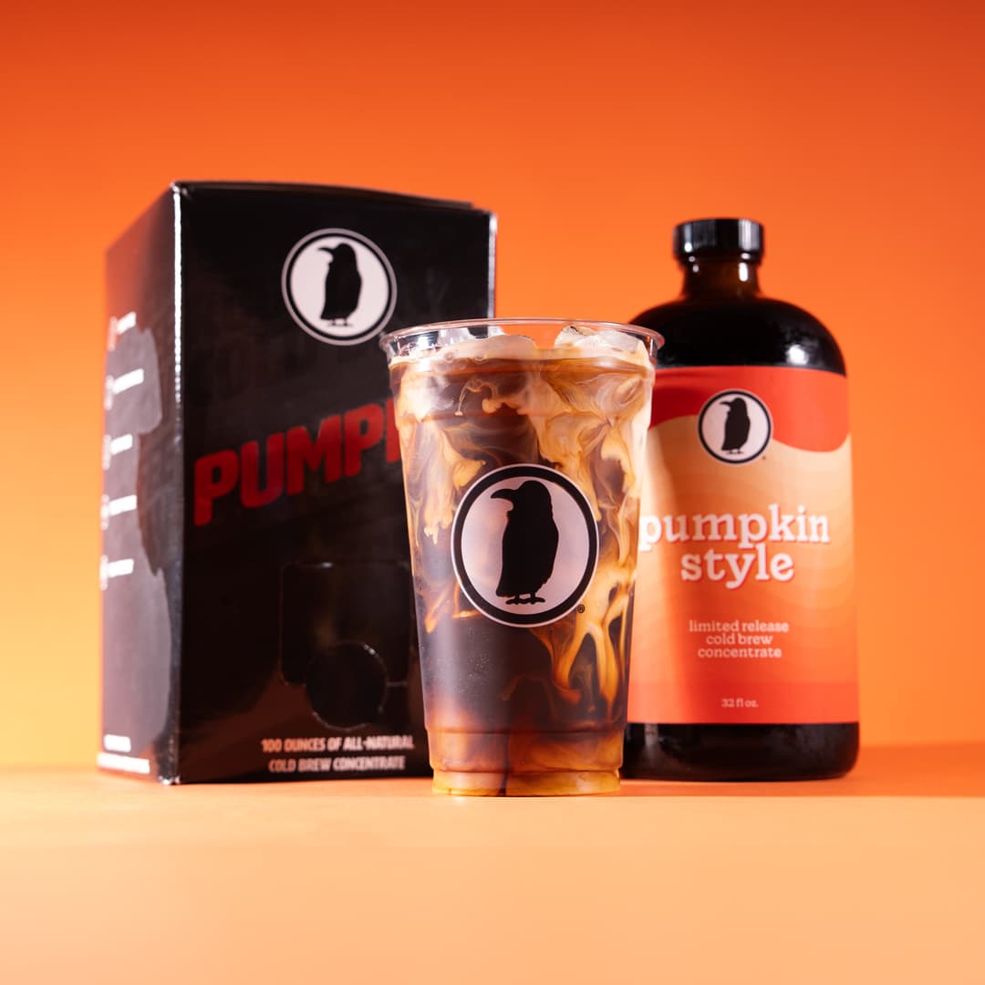 A bottle and box of Pumpkin Style cold brew concentrate behind a Rook cup filled with swirling cold brew, set against a solid orange background.