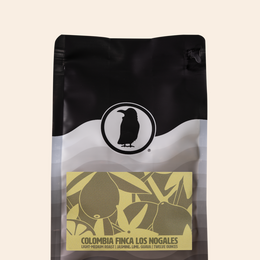 Bag of Colombia Finca Los Nogales coffee with a yellow label featuring abstract fruit and foliage illustrations. Text on the label reads: 'Light-Medium Roast | Jasmine, Lime, Guava | Twelve Ounces'.