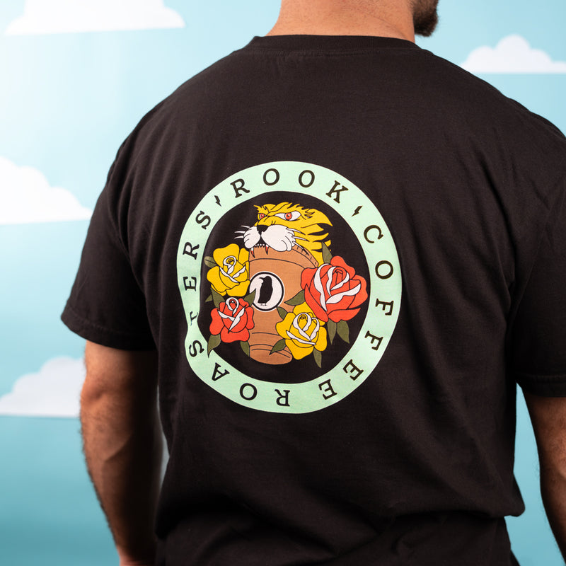 A man wearing a black tee shirt. The back of the shirt has a design, 'Rook Coffee Roasters' in a blue circle of text with a tiger clawing a cold brew cup in the center, surrounded by flowers. 