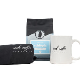 Folded black Rook Coffee T-shirt next to a white mug with the Rook Coffee Roasters logo and a bag of coffee.