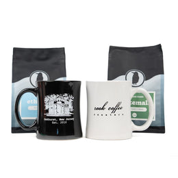 Two mugs, one black and one white, with Rook Coffee branding in front of two Rook Coffee bags.